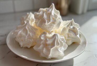 A close-up of several perfectly piped, white meringue cookies stacked on a small white plate.