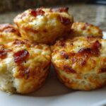 A close-up of several golden-brown Pizza Muffins topped with melted cheese and crispy bacon bits served on a white plate.