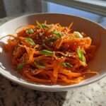 A close-up of a vibrant raw carrot salad, featuring julienned carrots topped with sesame seeds and sliced green onions.