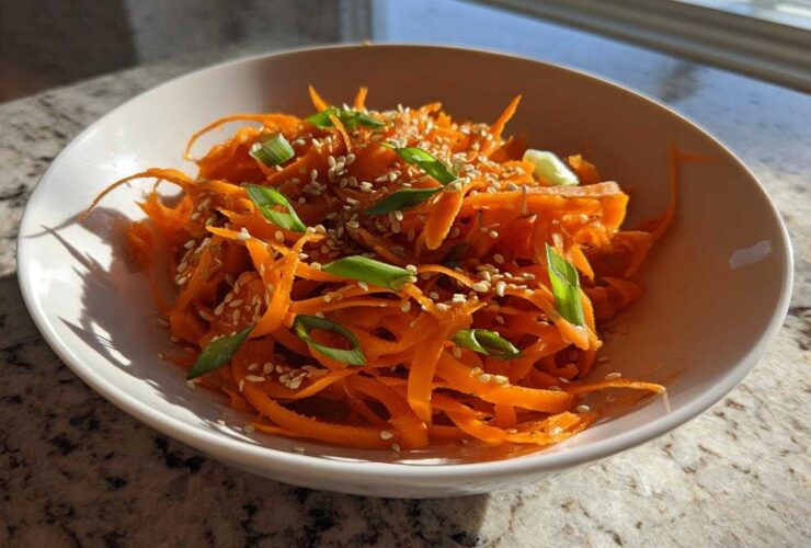 A close-up of a vibrant raw carrot salad, featuring julienned carrots topped with sesame seeds and sliced green onions.