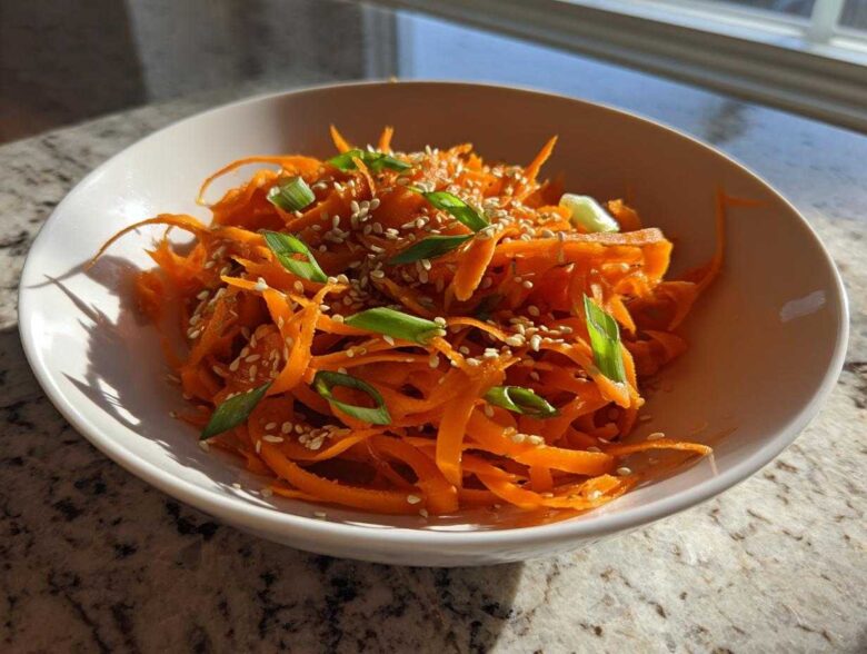 A close-up of a vibrant raw carrot salad, featuring julienned carrots topped with sesame seeds and sliced green onions.