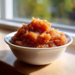 A close-up of chunky, dark brown applesauce piled high in a white bowl, sitting on a sunny windowsill.
