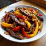 A close-up of sizzling steak fajitas featuring strips of seared steak mixed with sautéed red and yellow bell peppers and onions in a white bowl.