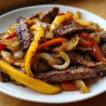 Close-up of sizzling steak fajitas featuring seasoned steak strips, yellow and red bell peppers, and caramelized onions on a white plate.
