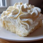 A close-up of a square slice of white cake generously topped with perfectly piped swirls of rich cream cheese frosting.