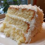 A close-up of a moist slice of coconut cake with white frosting and shredded coconut topping.