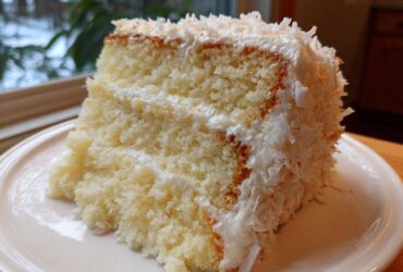 A close-up of a moist slice of coconut cake with white frosting and shredded coconut topping.