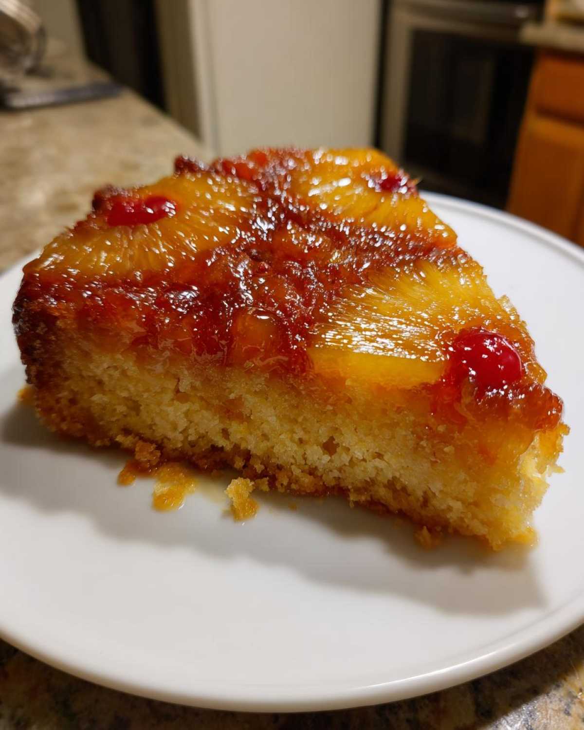 A close-up view of a moist slice of pineapple upside down cake with caramelized pineapple and cherries on a white plate.