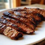 Slices of grilled steak showing a medium-rare center, coated in a rich Carne asada marinade, served on a white plate.