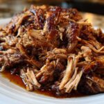 Close-up of a generous mound of saucy, shredded Slow Cooker Pulled Pork piled high on a white plate.