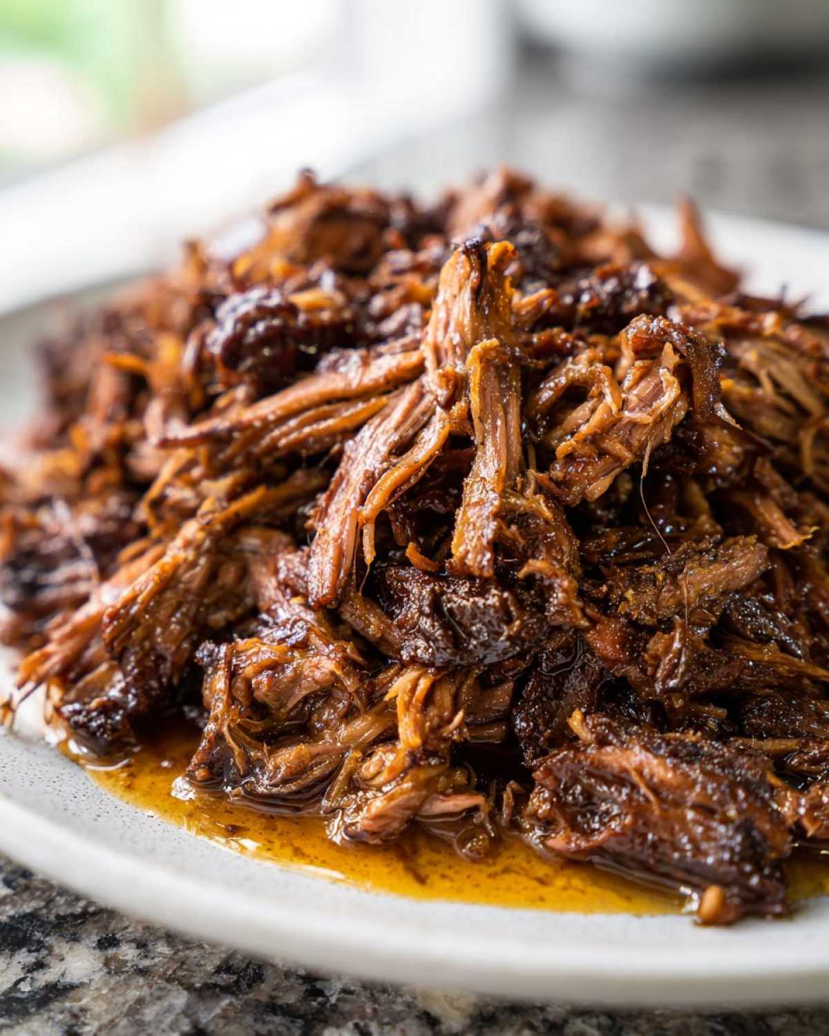 Close-up of juicy, shredded Slow Cooker Pulled Pork piled high on a light-colored plate with visible sauce.