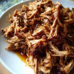 Close-up of tender, shredded Slow Cooker Pulled Pork glistening with sauce on a white plate.