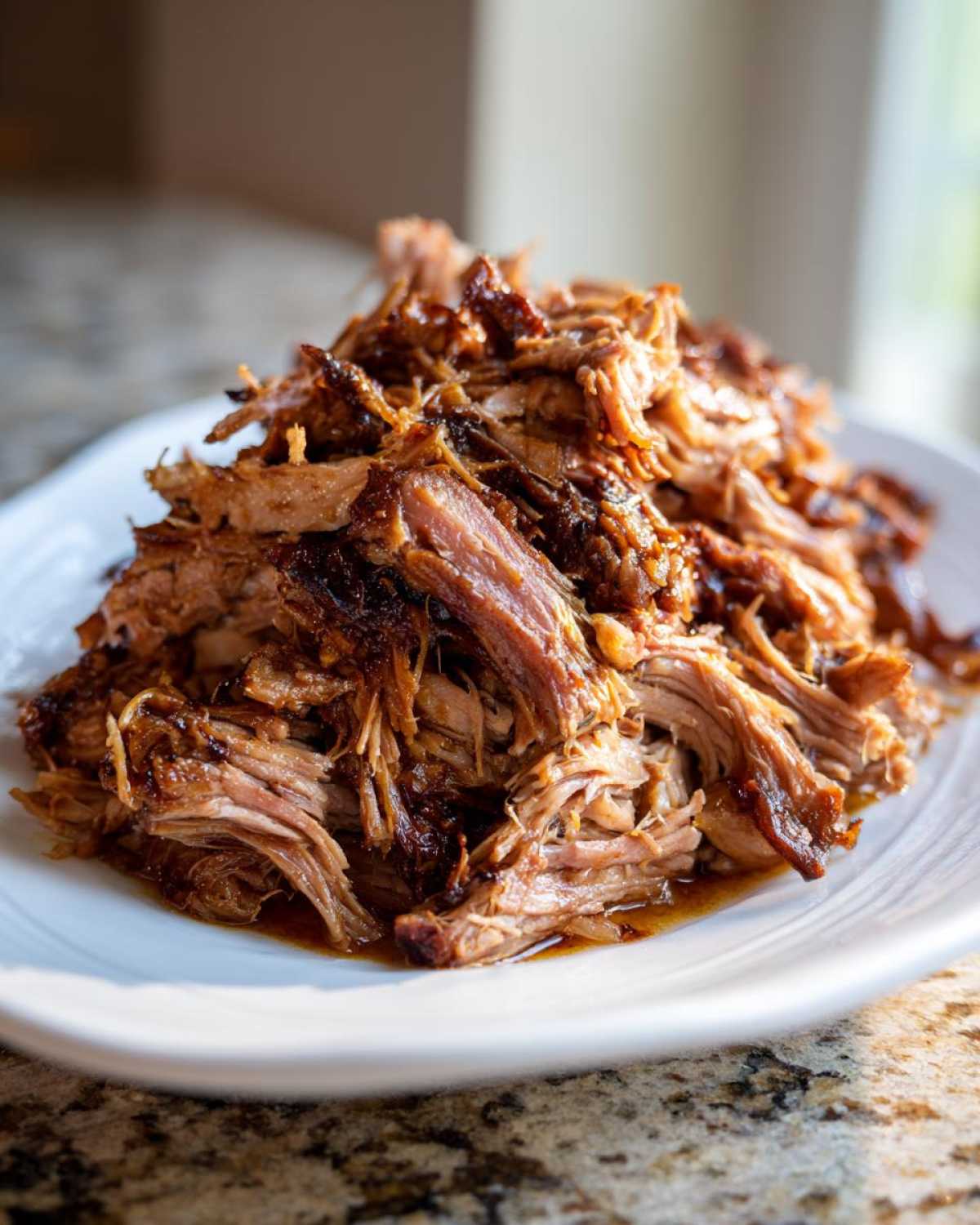 A close-up of a mound of moist, shredded Slow Cooker Pulled Pork glistening with sauce on a white plate.