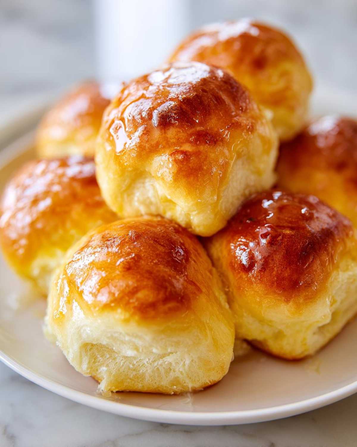 A stack of golden brown, glossy dinner rolls brushed with butter or glaze on a white plate.