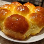 A cluster of freshly baked, golden brown dinner rolls with a shiny glaze arranged on a white plate.