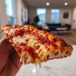 Close-up of a hand holding a slice of cheesy pizza made with sourdough discard pizza dough, showing airy crust.