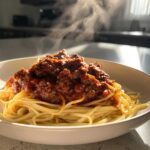Close-up of steaming spaghetti topped generously with rich bolognese sauce in a white bowl.