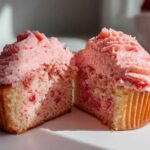 A strawberry cupcake cut in half showing the moist crumb with strawberry pieces and pink frosting.