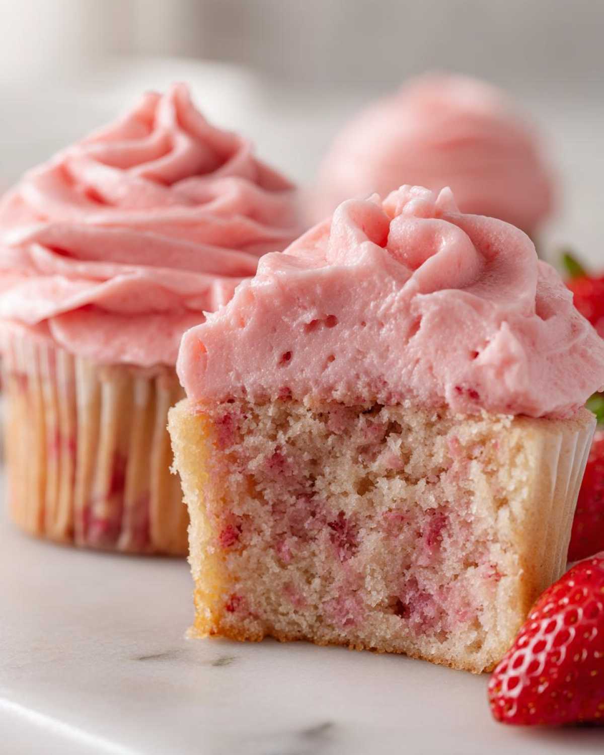 Close-up of a strawberry cupcake cut in half showing the moist cake filled with strawberry pieces, topped with pink frosting.