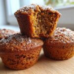 A stack of moist, brown bran muffins topped with coarse sugar crystals, one muffin is broken open.