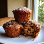 Three delicious bran muffins stacked on a white plate, topped with coarse sugar; one muffin has a bite taken out showing the texture.