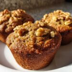 Three freshly baked banana nut muffins, golden brown and topped with crunchy walnuts, sitting on a white plate.