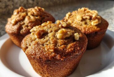 Three freshly baked banana nut muffins, golden brown and topped with crunchy walnuts, sitting on a white plate.
