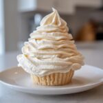 A single cupcake topped with a tall swirl of creamy vanilla buttercream frosting on a white plate.