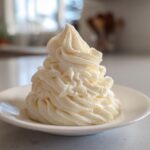 A tall, perfectly piped swirl of creamy vanilla buttercream frosting on a small white saucer.