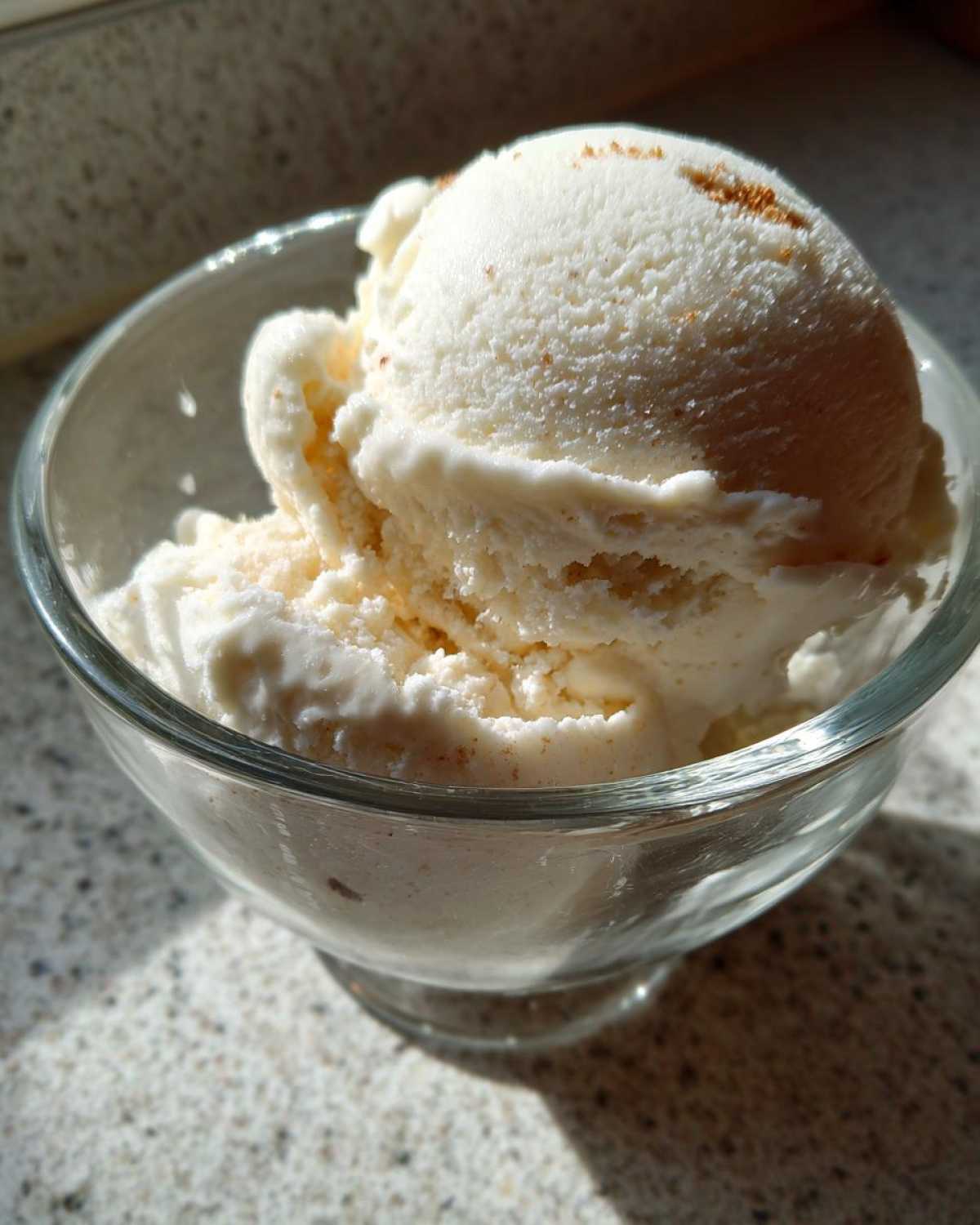 A close-up of a scoop of creamy white protein ice cream in a glass bowl, lightly dusted with cinnamon.