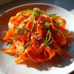 Close-up of a vibrant raw carrot salad made with shaved carrot ribbons, topped with sesame seeds and green onions.