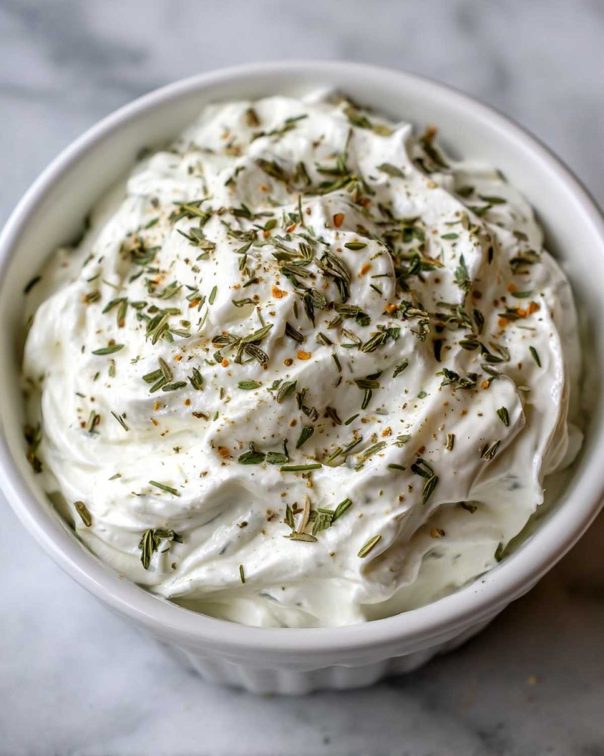 A close-up of thick, creamy 5-Minute Greek Yogurt Ranch Dip topped with dried herbs and spices in a white bowl.