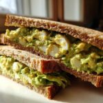 Two halves of an Avocado Egg Salad Sandwich stacked, showing creamy green filling and sliced hard-boiled eggs on toasted whole wheat bread.