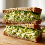 Two halves of an Avocado Egg Salad Sandwich stacked, showing creamy green filling with egg chunks on whole wheat bread.
