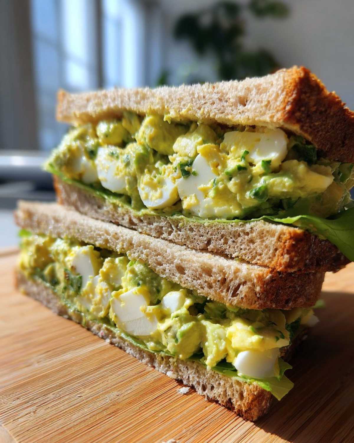 Two halves of an Avocado Egg Salad Sandwich stacked on a wooden board, showing creamy filling and hard-boiled egg chunks.