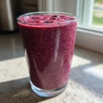 A tall glass filled with a thick, deep purple Blueberry Lemon Chia Smoothie sitting on a sunlit countertop.