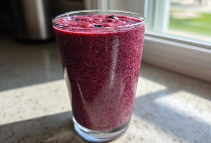 A tall glass filled with a thick, deep purple Blueberry Lemon Chia Smoothie sitting on a sunlit countertop.
