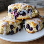 Three delicious Blueberry Lemon Scones stacked on a white plate, generously drizzled with white lemon glaze.