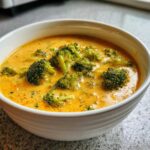 Close-up of a creamy, orange-yellow Copycat Broccoli Cheddar Soup filled with bright green broccoli florets in a white bowl.