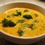 Close-up of a creamy, orange-yellow Copycat Broccoli Cheddar Soup filled with bright green broccoli florets in a white bowl.