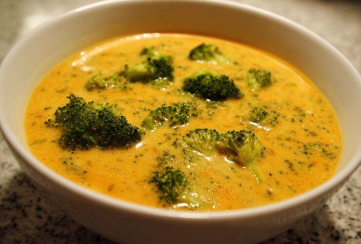 Close-up of a creamy, orange-yellow Copycat Broccoli Cheddar Soup filled with bright green broccoli florets in a white bowl.