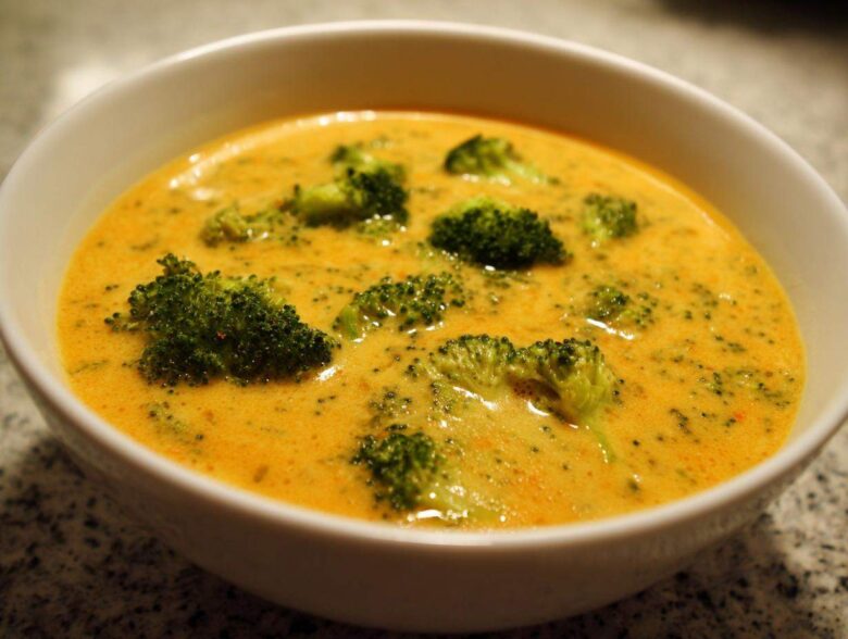 Close-up of a creamy, orange-yellow Copycat Broccoli Cheddar Soup filled with bright green broccoli florets in a white bowl.