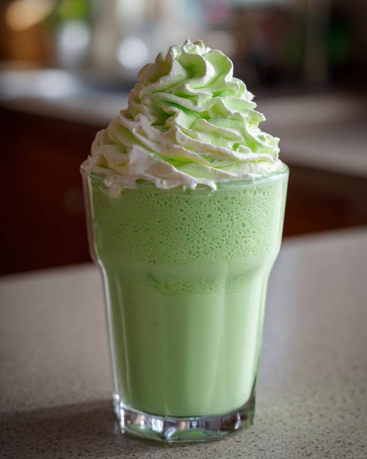 A tall glass filled with a vibrant green Copycat Shamrock Shake, topped high with swirls of white and light green mint whipped cream.
