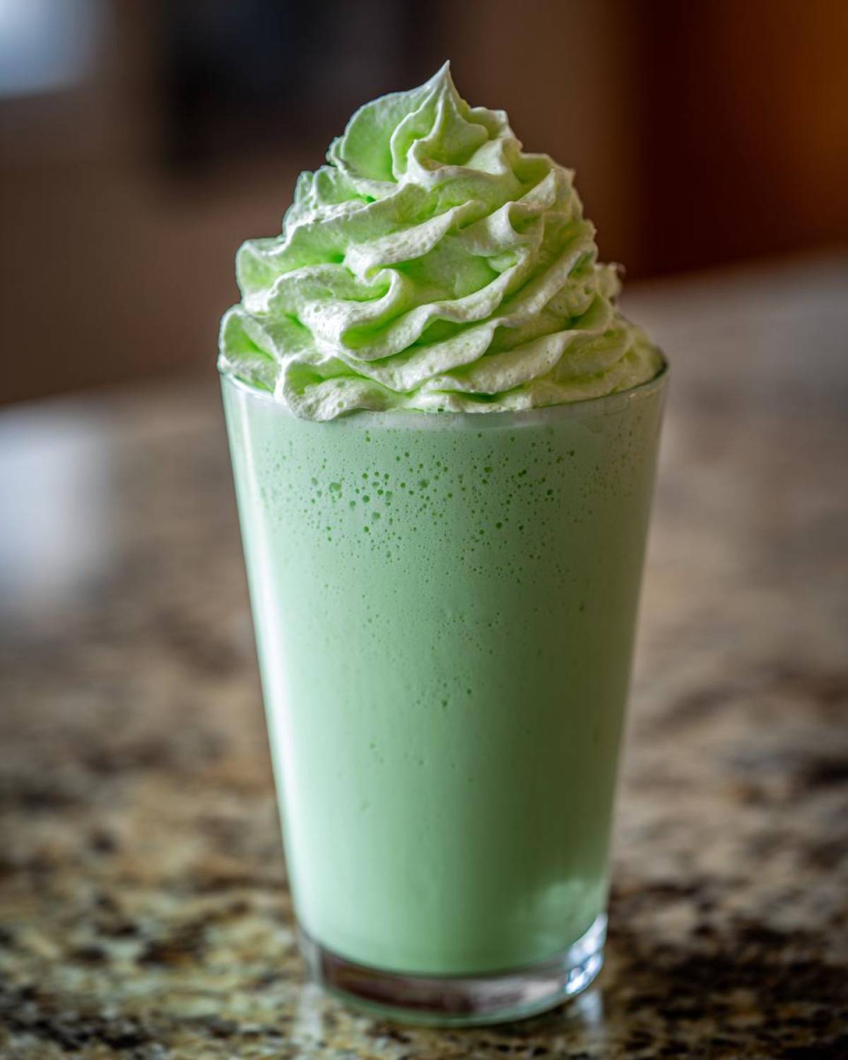 A tall glass filled with a vibrant green Copycat Shamrock Shake topped with a swirl of light green mint whipped cream.