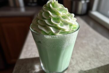 A tall glass of bright green Copycat Shamrock Shake topped with a swirl of mint whipped cream.