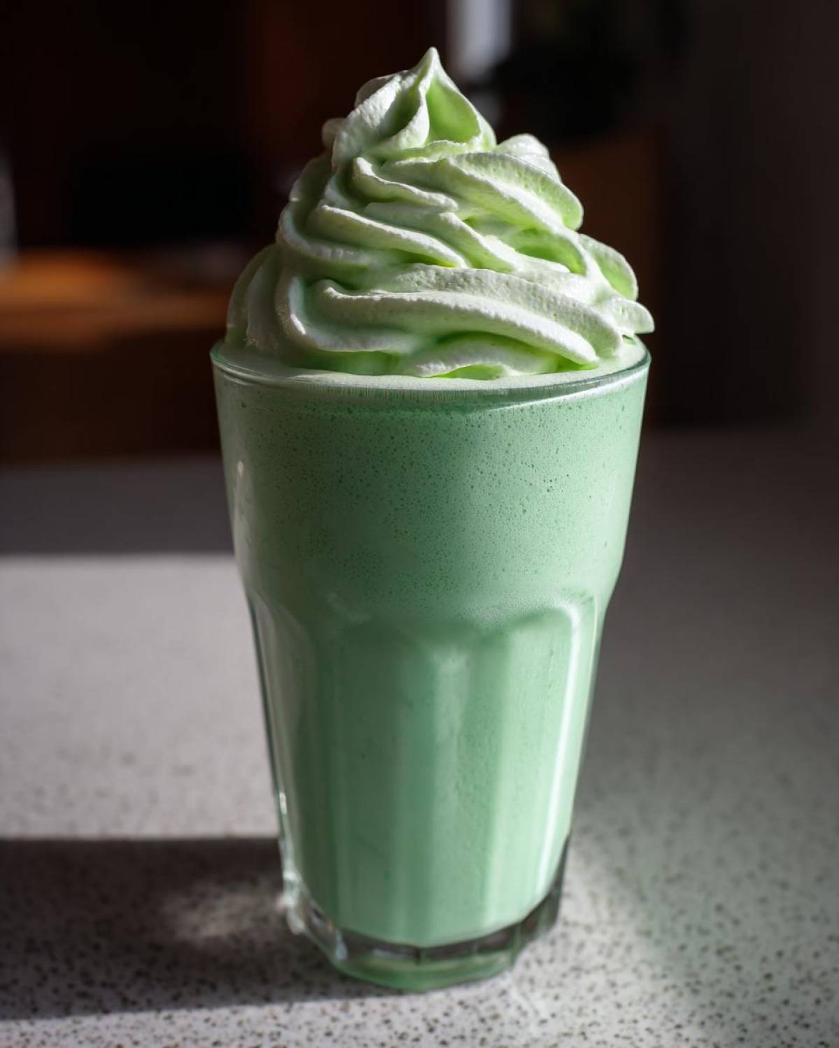 A tall glass filled with a vibrant green Copycat Shamrock Shake topped with a swirl of mint whipped cream.