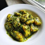 Close-up of plump, yellow gnocchi generously coated in vibrant green, creamy pesto sauce, served in a white bowl.