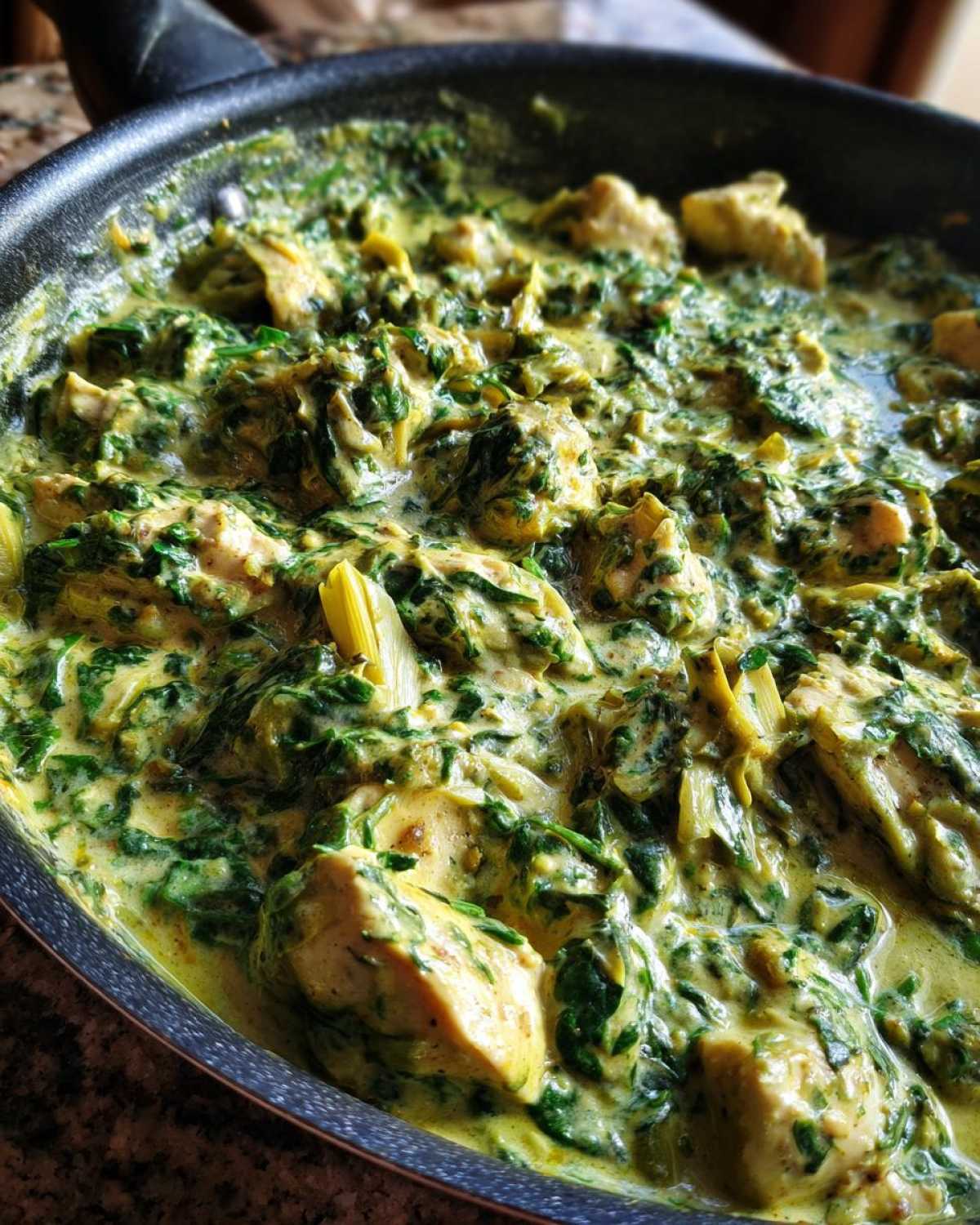 Close-up of chunks of chicken simmering in a rich, creamy sauce with spinach and artichoke hearts in a skillet.