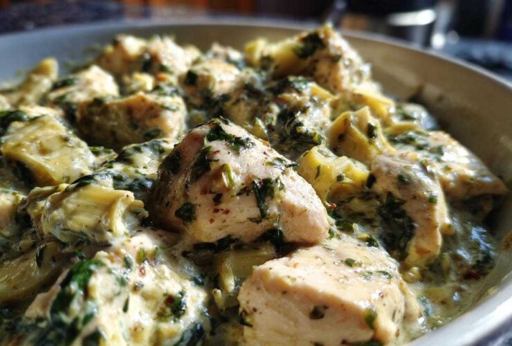 Close-up of tender chicken pieces coated in a rich, creamy sauce with spinach and artichoke hearts in a skillet.