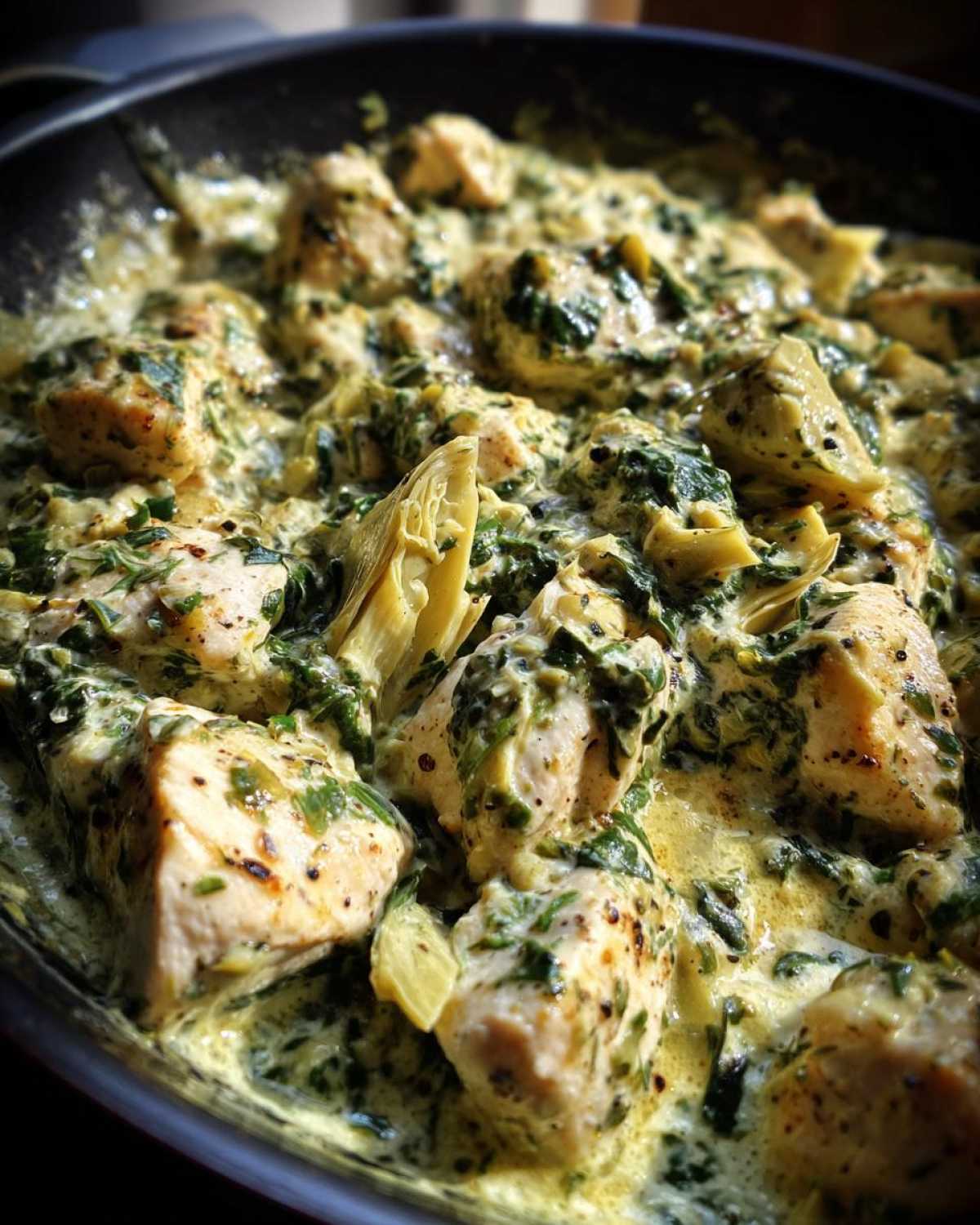 Close-up of chunks of chicken simmering in a rich, creamy spinach artichoke sauce in a black skillet.